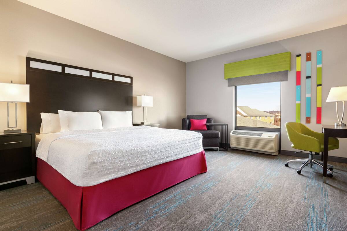 A hotel room with a large bed, a desk, a chair, and a window with a city view.