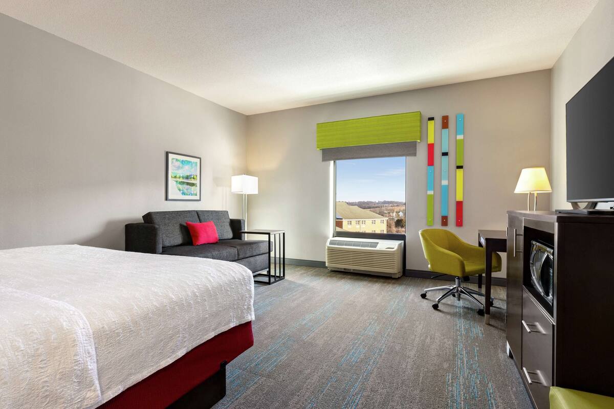 A hotel room with a bed, a sofa, a desk, a chair, a TV, and a window with a view.