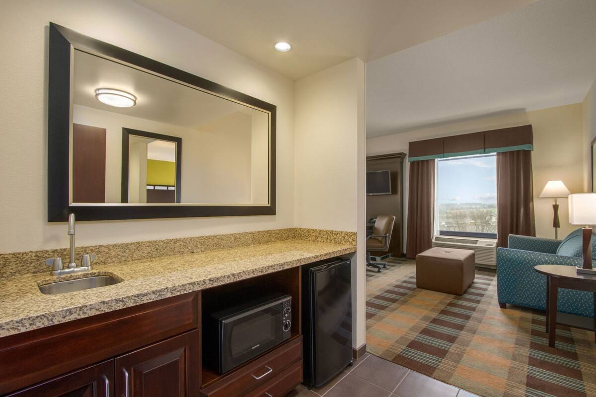 A hotel room with a kitchenette, a large mirror, a TV, a sofa, and a window with curtains.