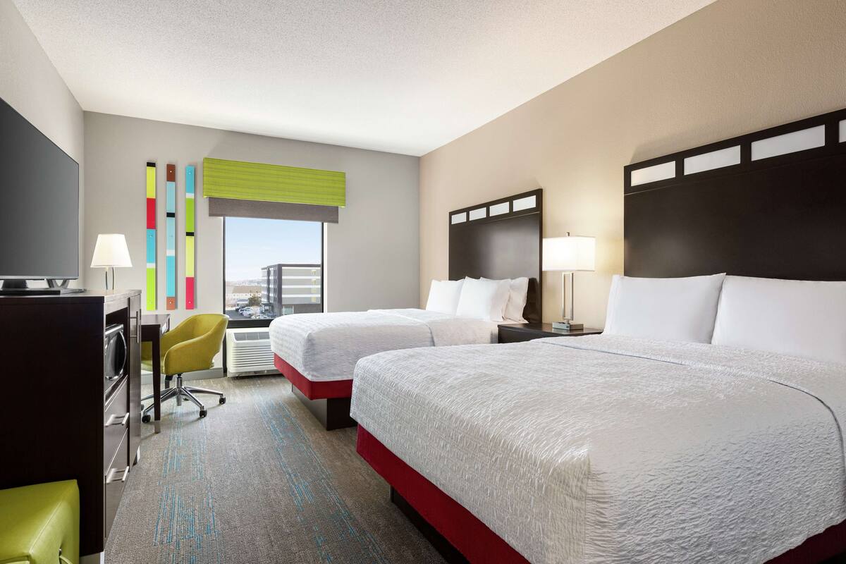 A hotel room with two beds, a desk, a chair, and a TV.