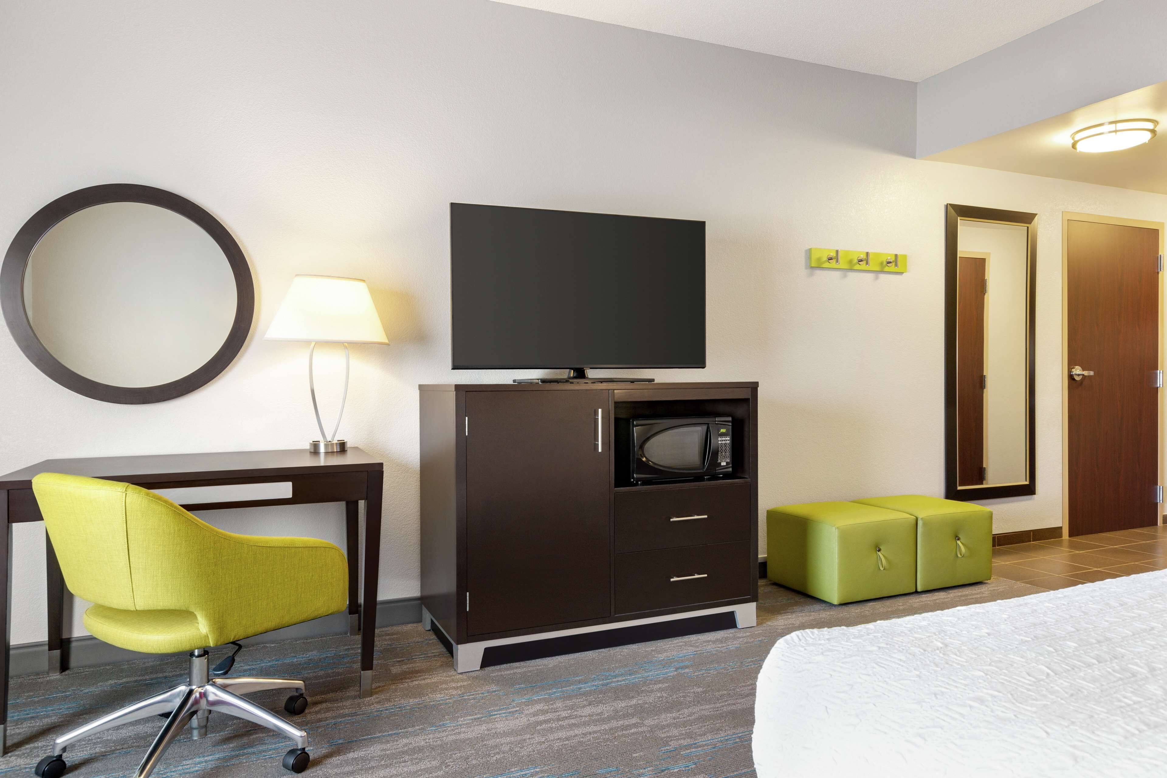 Quarto de hotel com escrivaninha, televisão, micro-ondas, um banquinho verde, espelho redondo e uma cama.