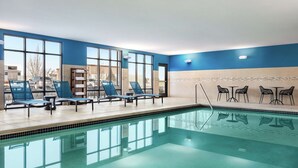 An indoor swimming pool with clear blue water, surrounded by lounge chairs and tables, large windows, and a view of the outside. - Hampton Inn & Suites York South (York)
