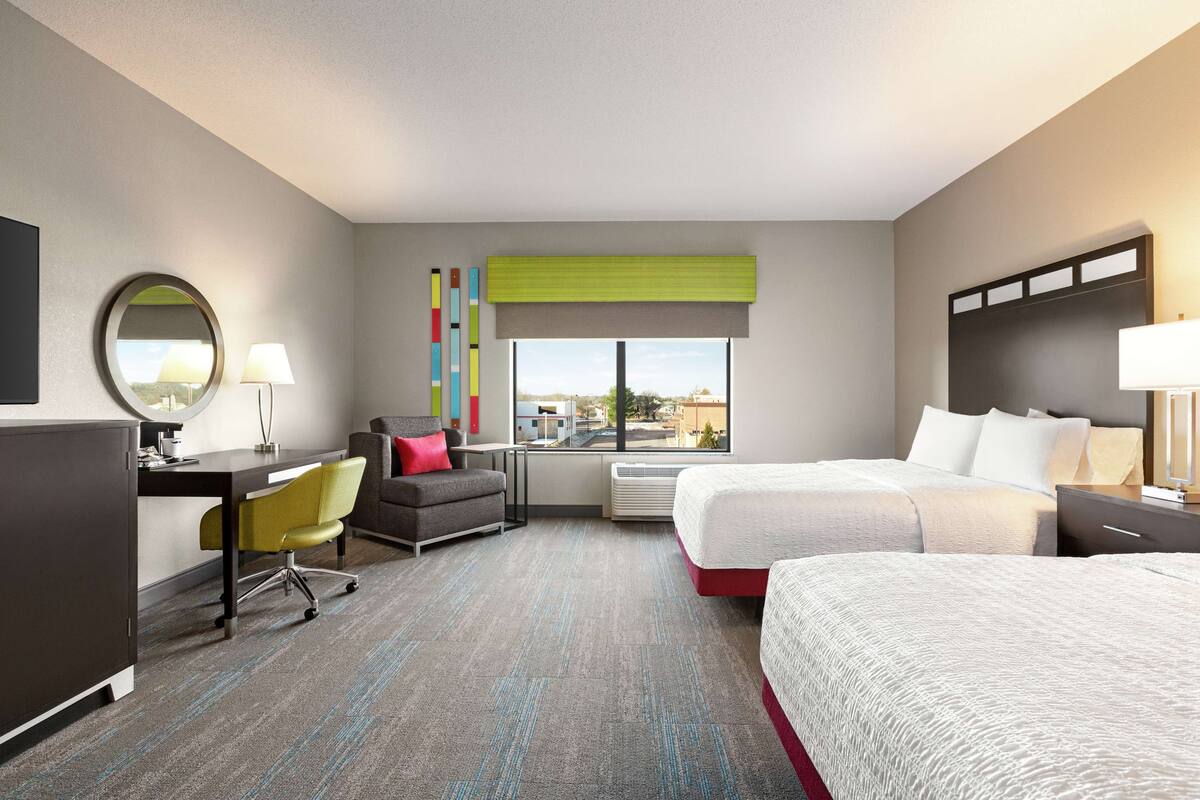 A hotel room with a desk, two beds, a chair, a window with a view, and a mirror.