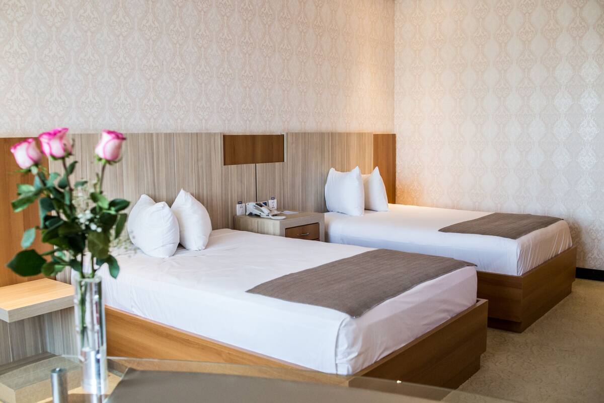 A hotel room with two beds, a nightstand, a lamp, and a vase with flowers.