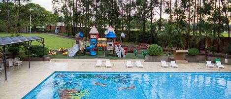 Una piscina con un diseño de mosaico colorido, rodeada de sillones de descanso y una zona de juegos infantiles con toboganes y estructuras para escalar.