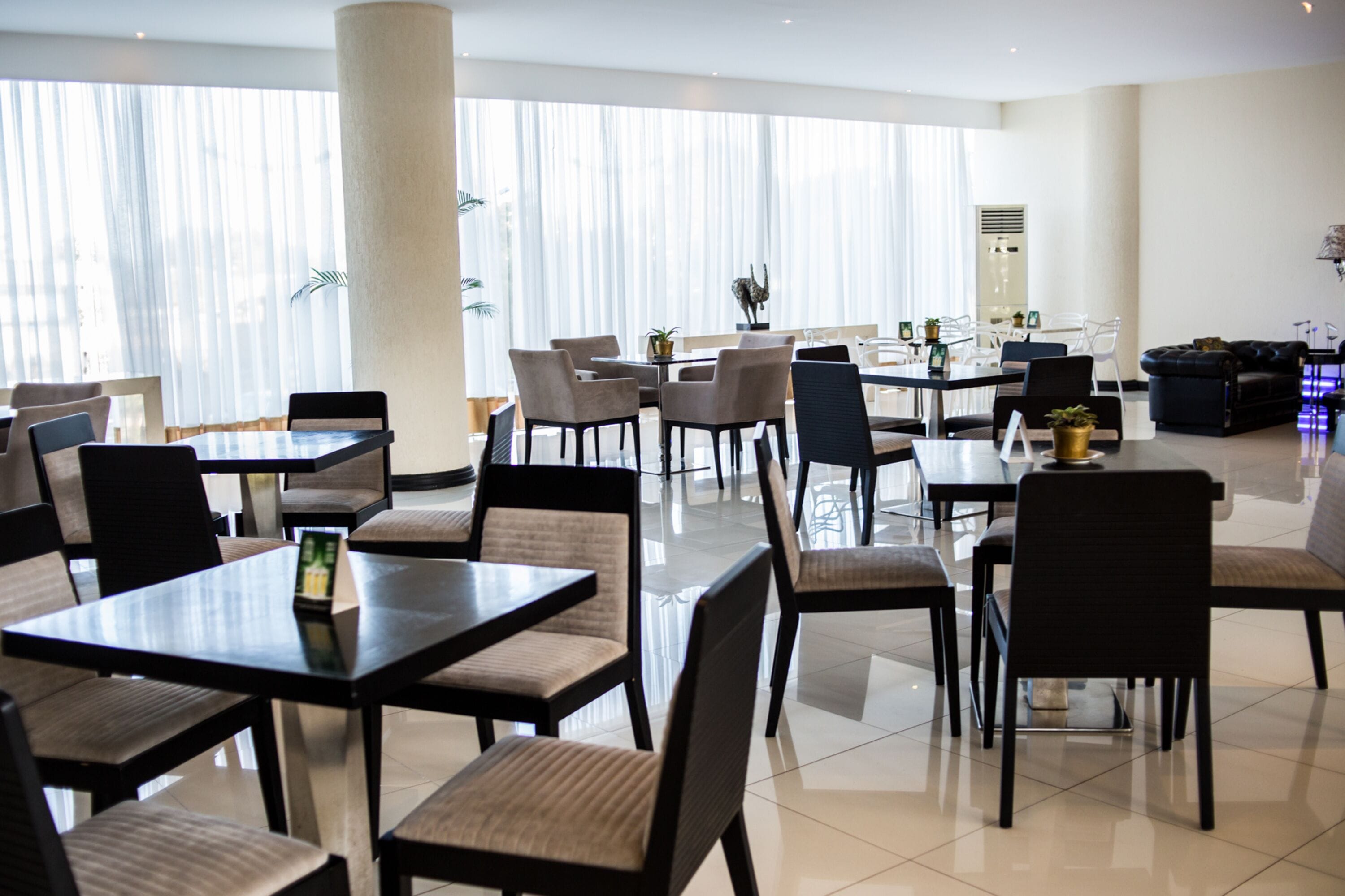 Een ruime hotel eetzaal met meerdere tafels en stoelen, grote ramen met lichte gordijnen en een modern interieur.