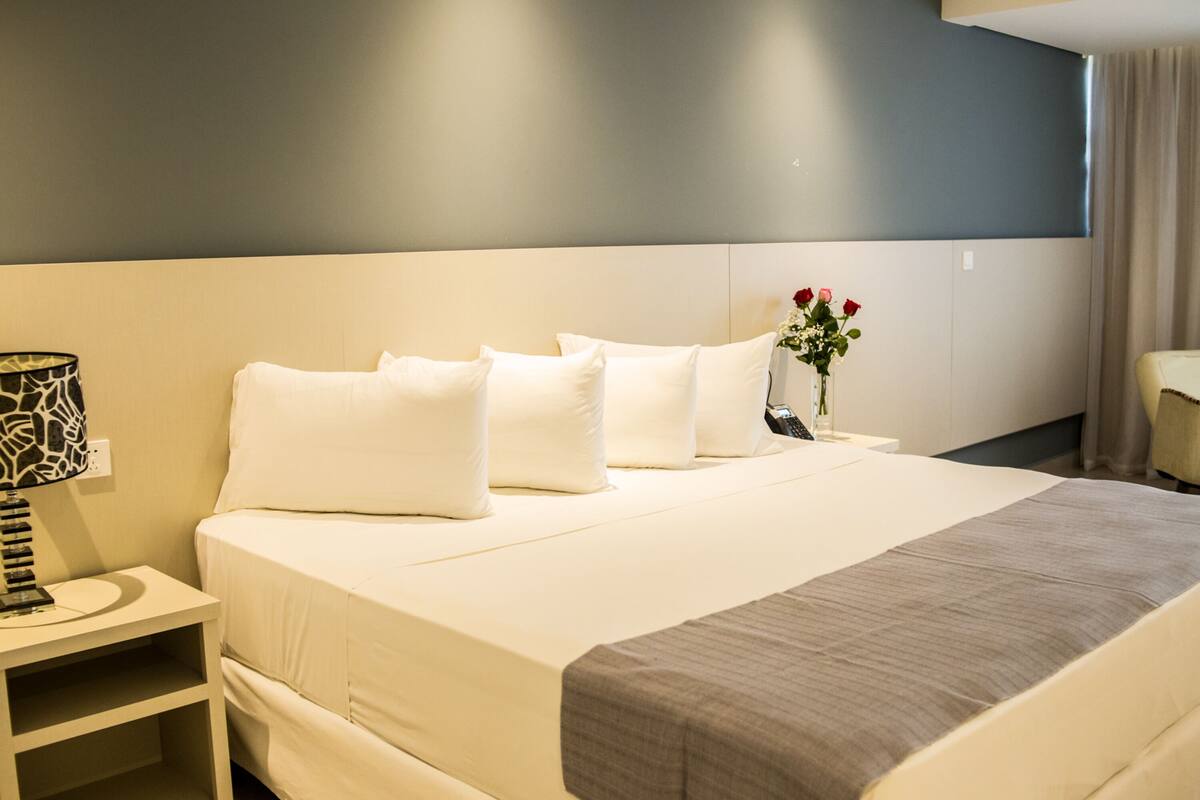 A neatly made bed with white linens and a gray throw blanket, a bedside table with a lamp, and a vase with red flowers.