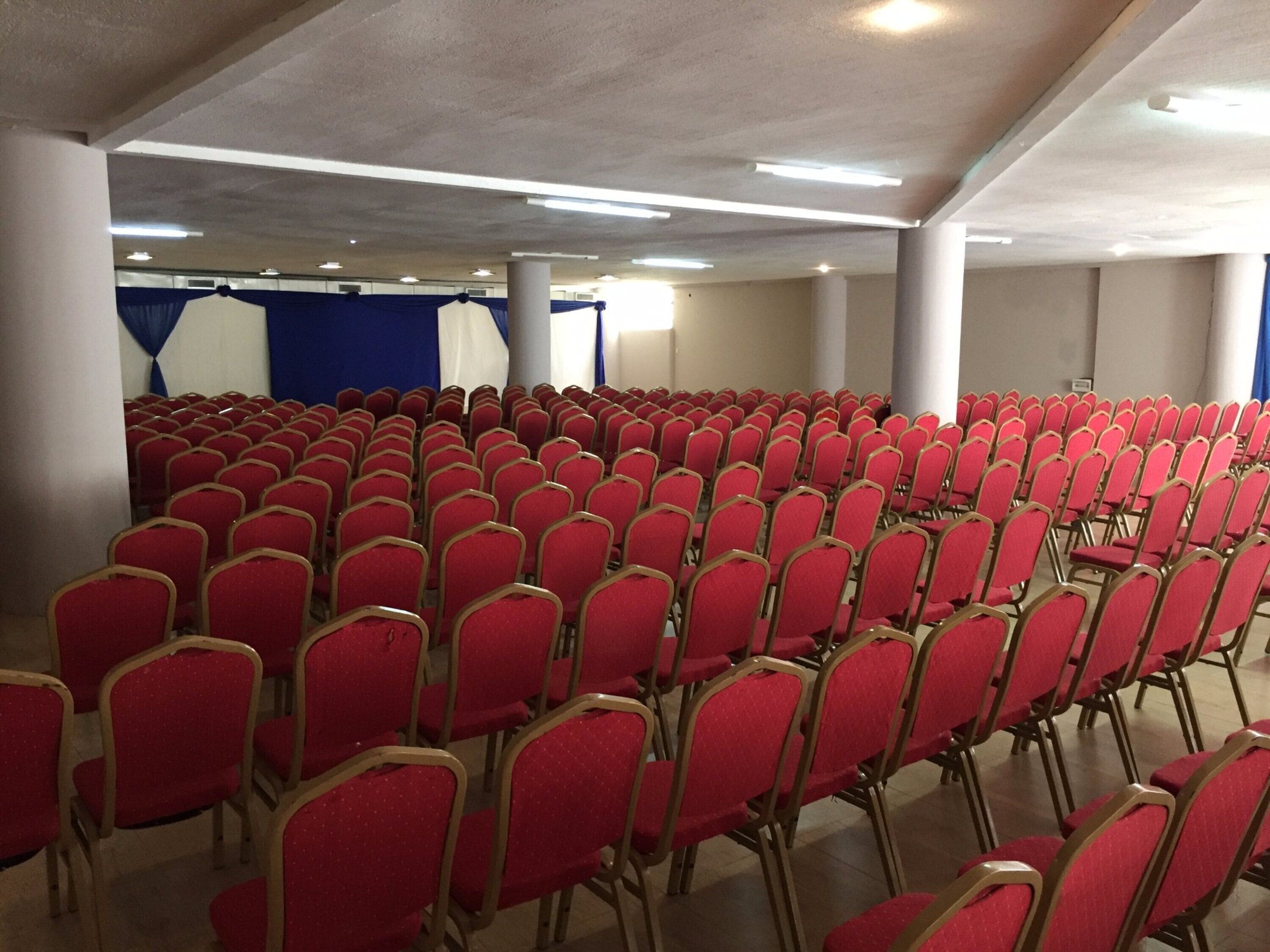 Um salão amplo com fileiras de cadeiras vermelhas e um palco com cortinas azuis.