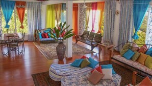 Amplia sala de estar con piso de madera, un conjunto de sofá, una mesa de comedor con sillas y un gran ventanal con cortinas coloridas.