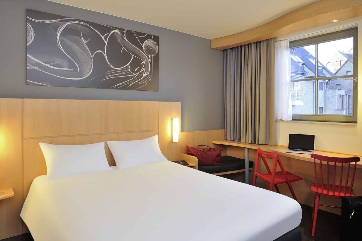 Een hotelkamer met een bed, een bureau, een stoel, een laptop, een rode tas en een raam met gordijnen.