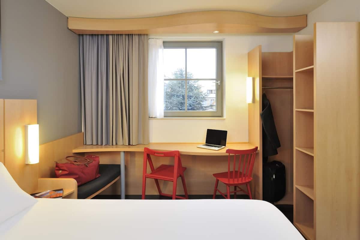 Une chambre dâhĂŽtel comprenant un lit, un bureau avec un ordinateur portable, deux chaises rouges, un porte-manteau et une armoire.