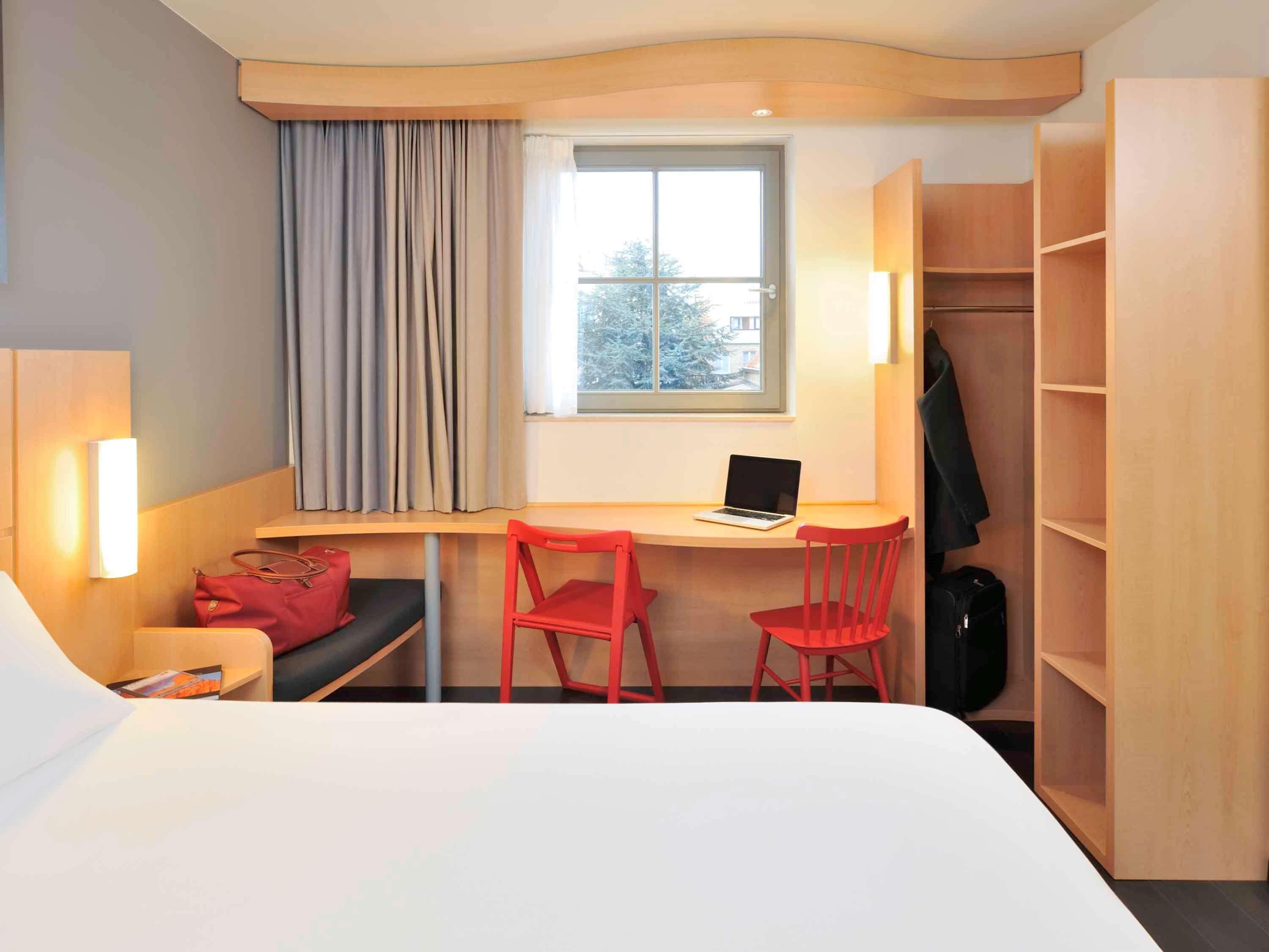 A hotel room with a bed, a desk with a laptop, two red chairs, a coat rack, and a wardrobe.
