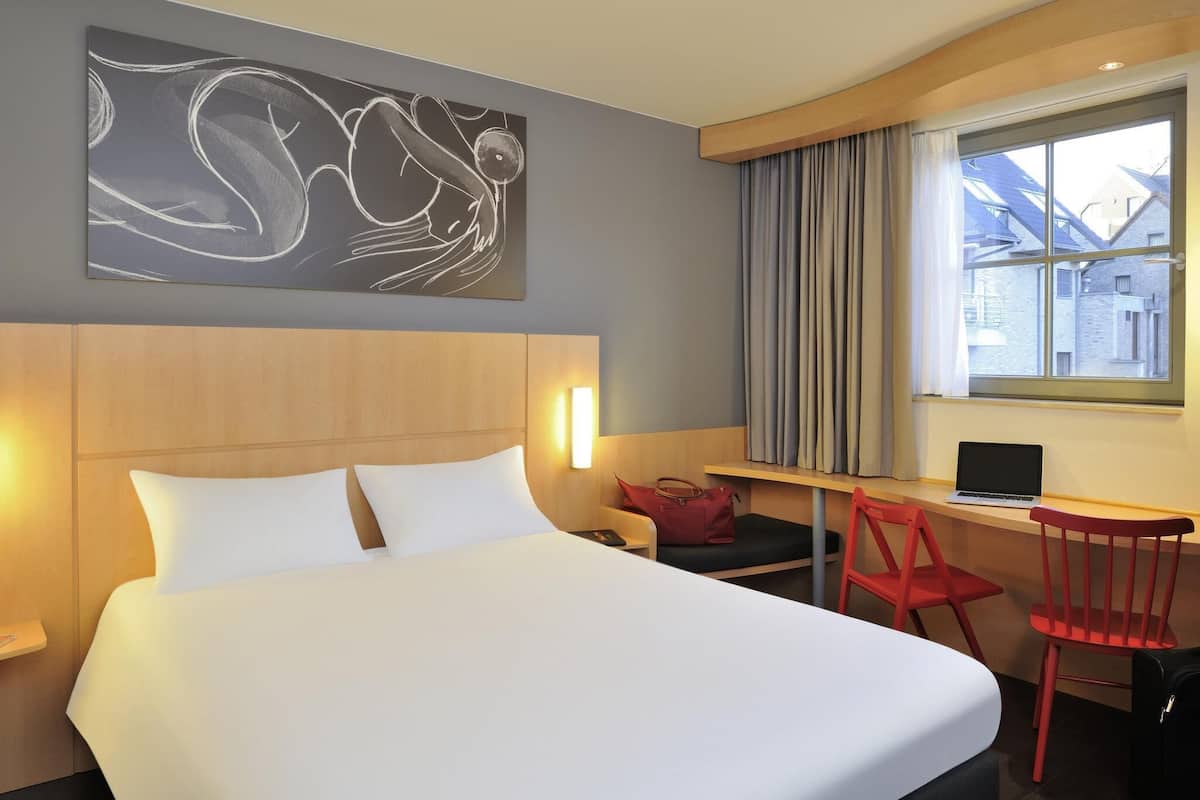 A hotel room with a bed, a desk, a chair, a laptop, and a window with curtains.
