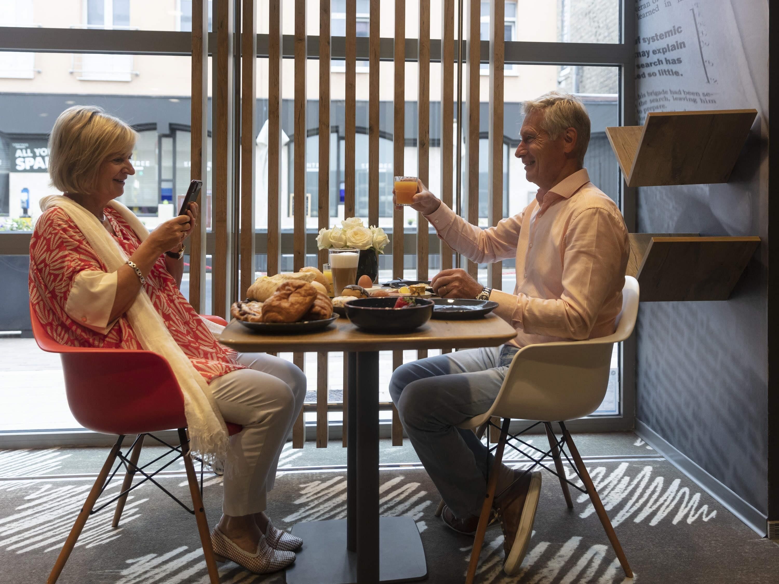 Two people seated at a table with food and drinks, in a room with large windows and a modern interior design.