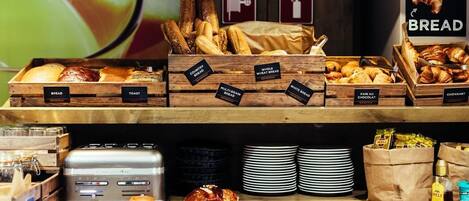 Una vitrina de panadería con distintos tipos de pan y pasteles, una tostadora y pilas de platos.