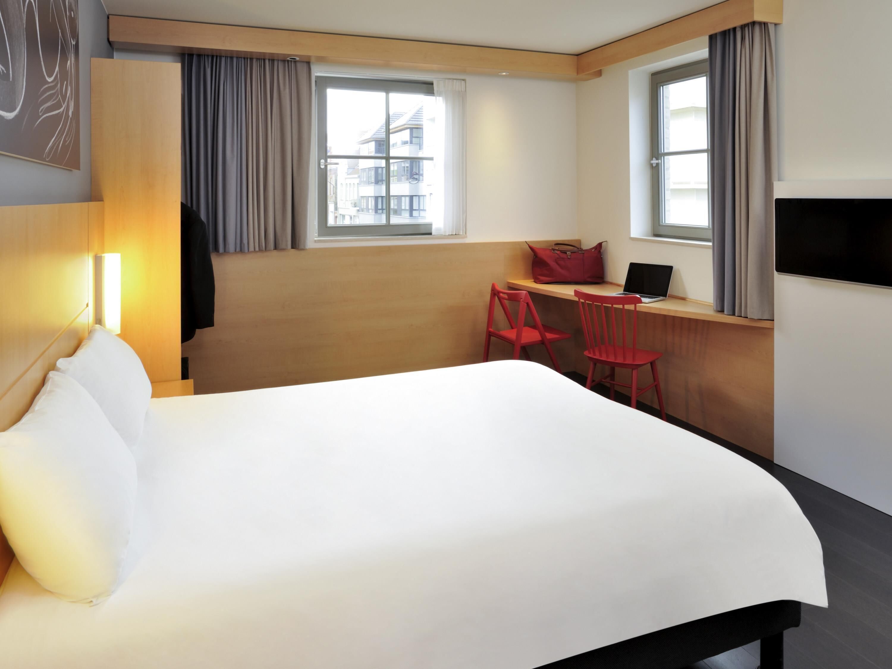 A hotel room with a bed, a desk with a laptop, a red chair, and a TV.