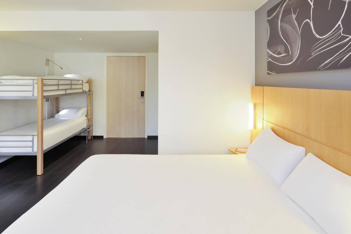 Een hotelkamer met een stapelbed, een enkel bed, een houten hoofdeinde, een nachtkastje en een kunstwerk aan de muur.