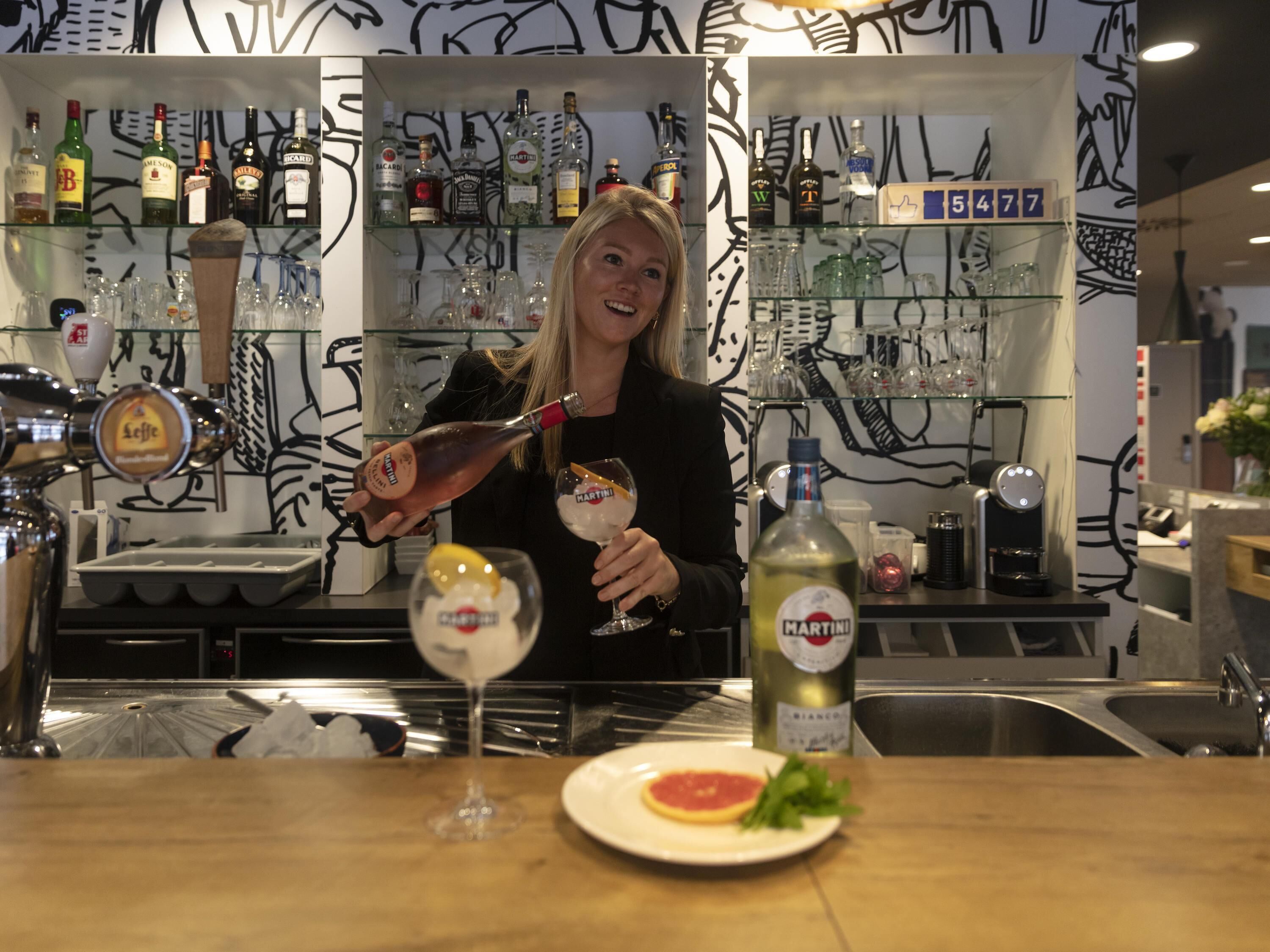 A bar area with a bartender pouring a drink into a glass, a bottle of Martini Martini on the counter, and a plate with a slice of lemon and a sprig of parsley.