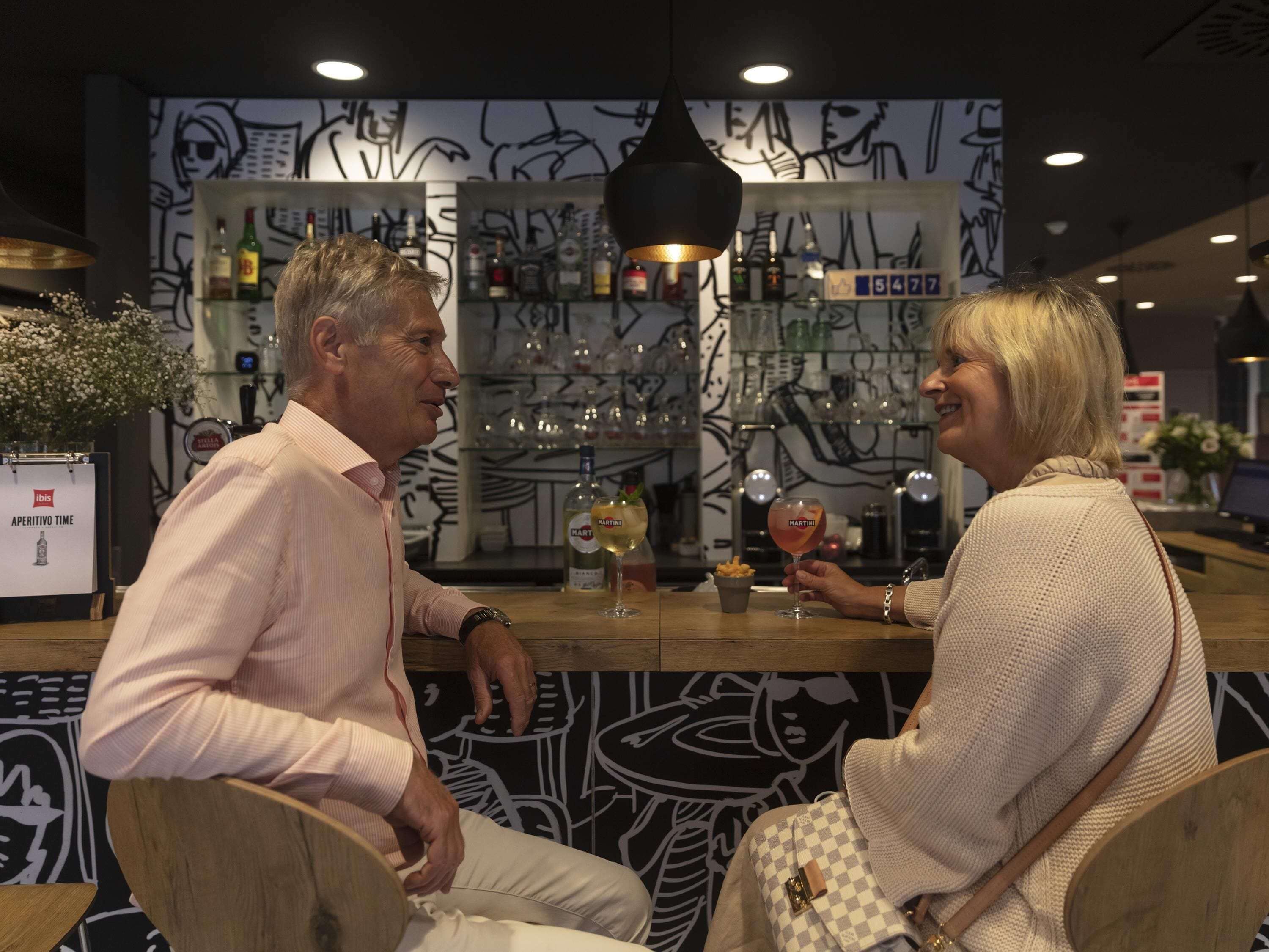 Twee personen zitten aan een bar met een drankje voor zich, een muur met abstracte kunst en planken met flessen.