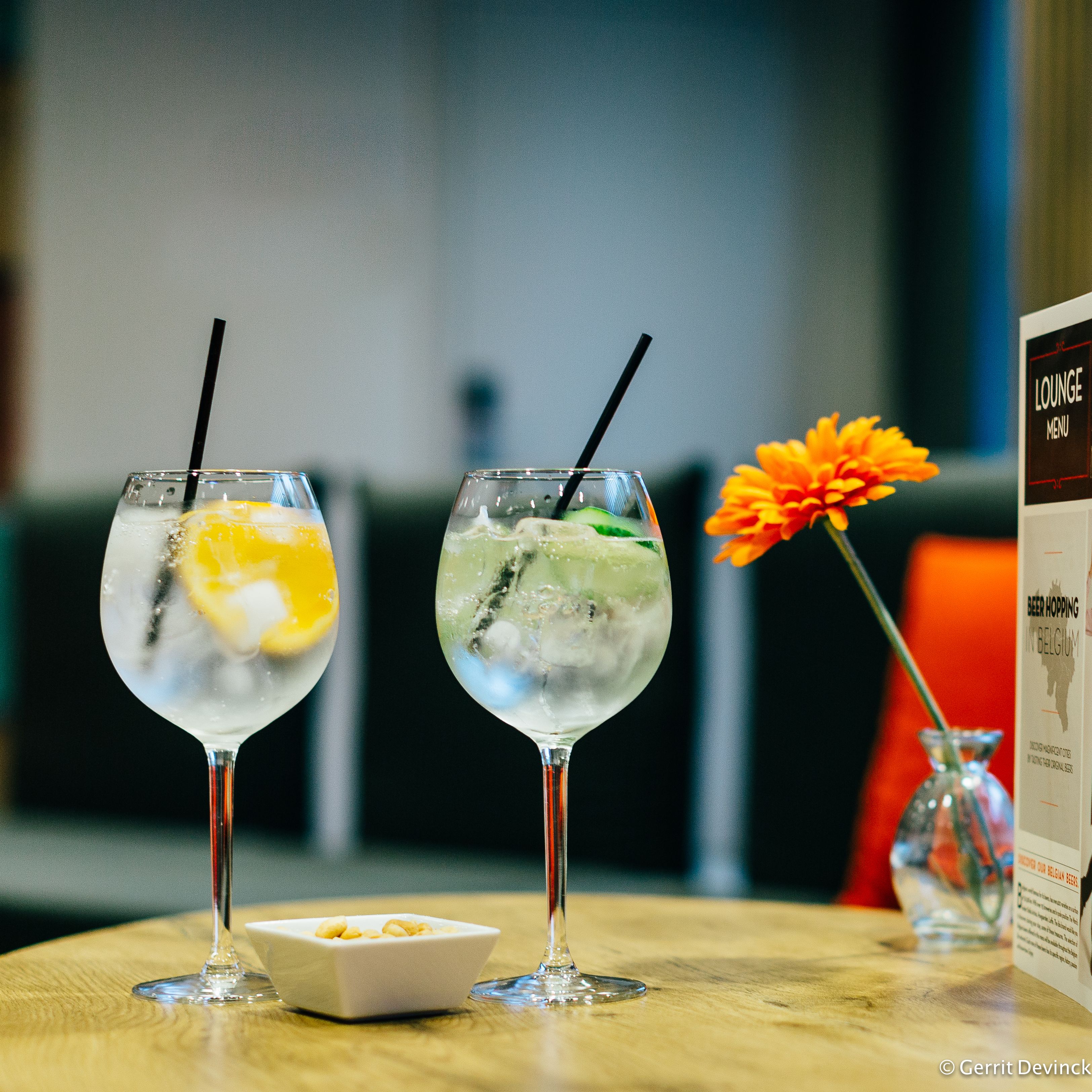 Two cocktails with straws on a table, a small bowl of nuts, and a menu stand with a flower.
