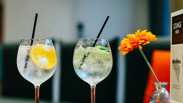 Two cocktails with straws on a table, a small bowl of nuts, and a menu stand with a flower.