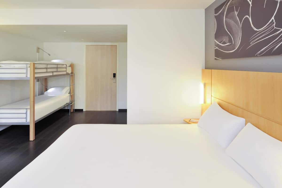 A hotel room with a bunk bed, a large bed, a wooden headboard, a bedside table, and a wall-mounted artwork.