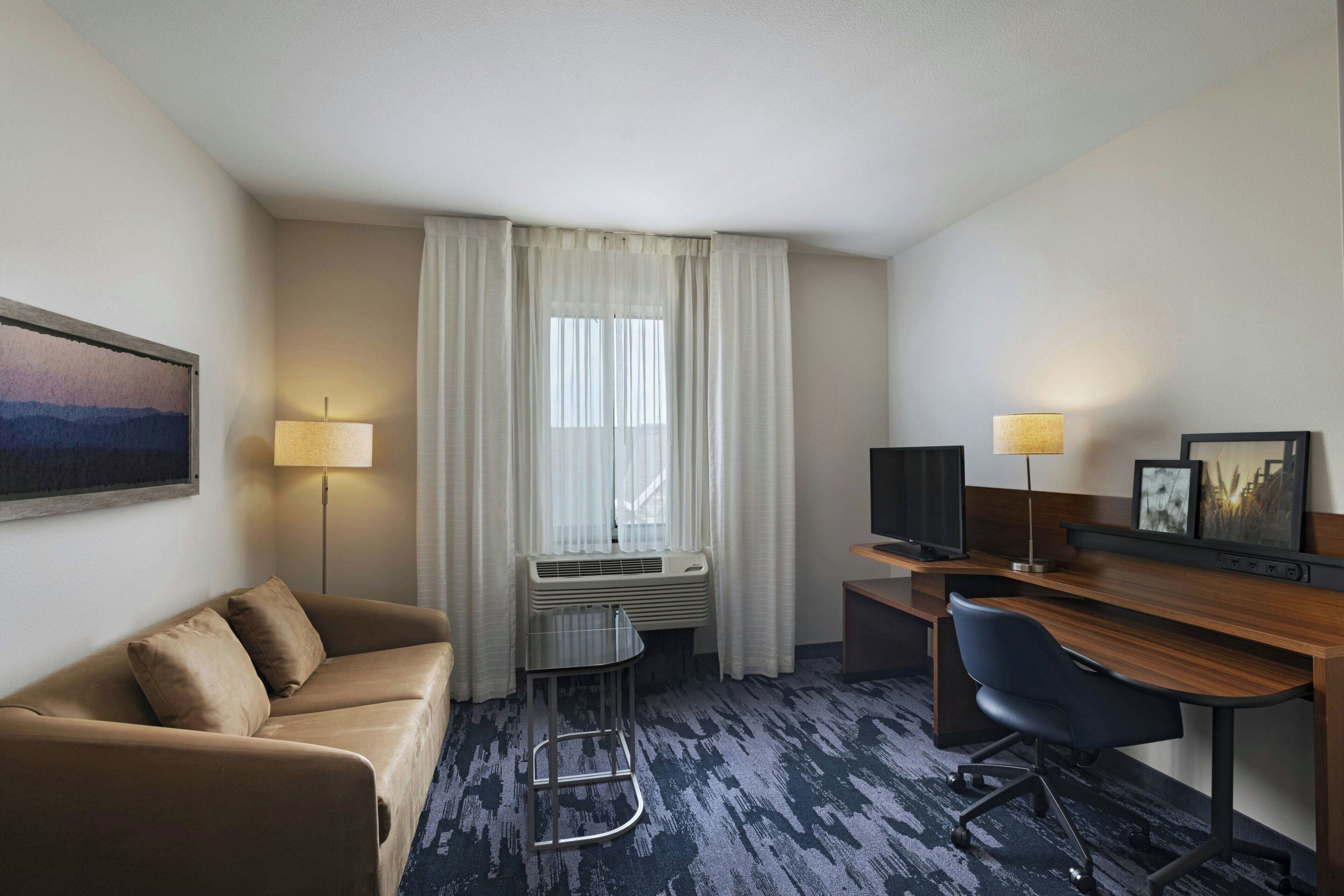 Hotelkamer met een bank, bureau, stoel, televisie, lamp en raam met gordijnen.