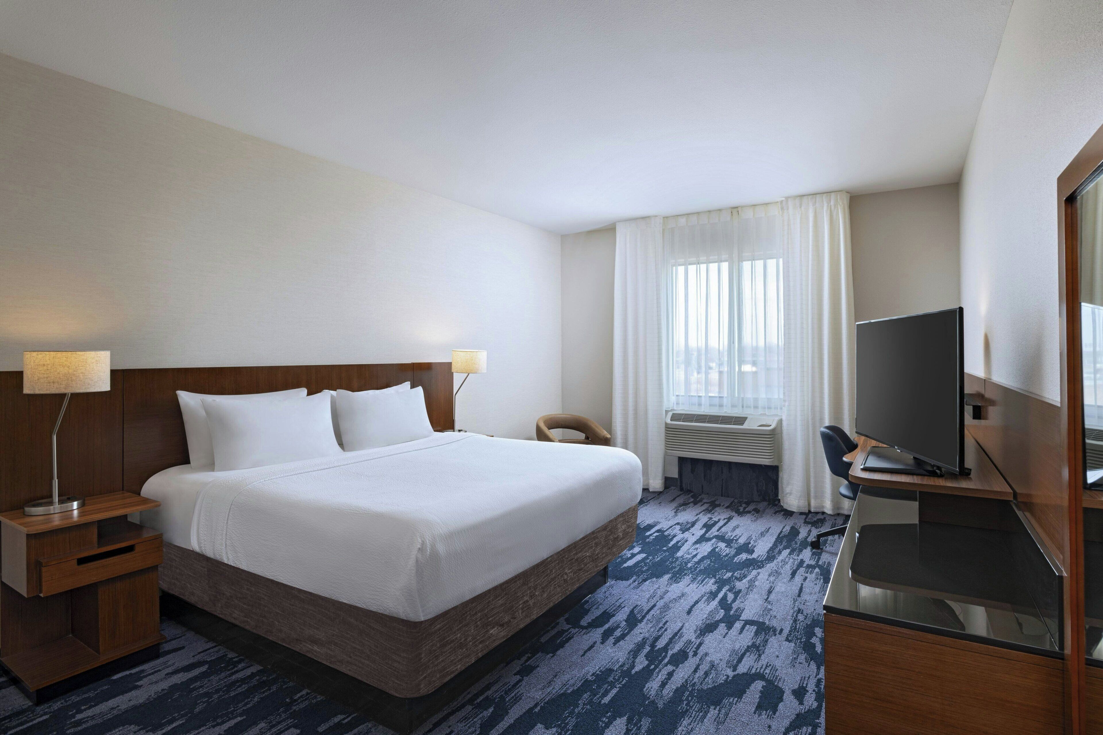 A hotel room with a large bed, a desk with a TV, a chair, and a window with curtains.