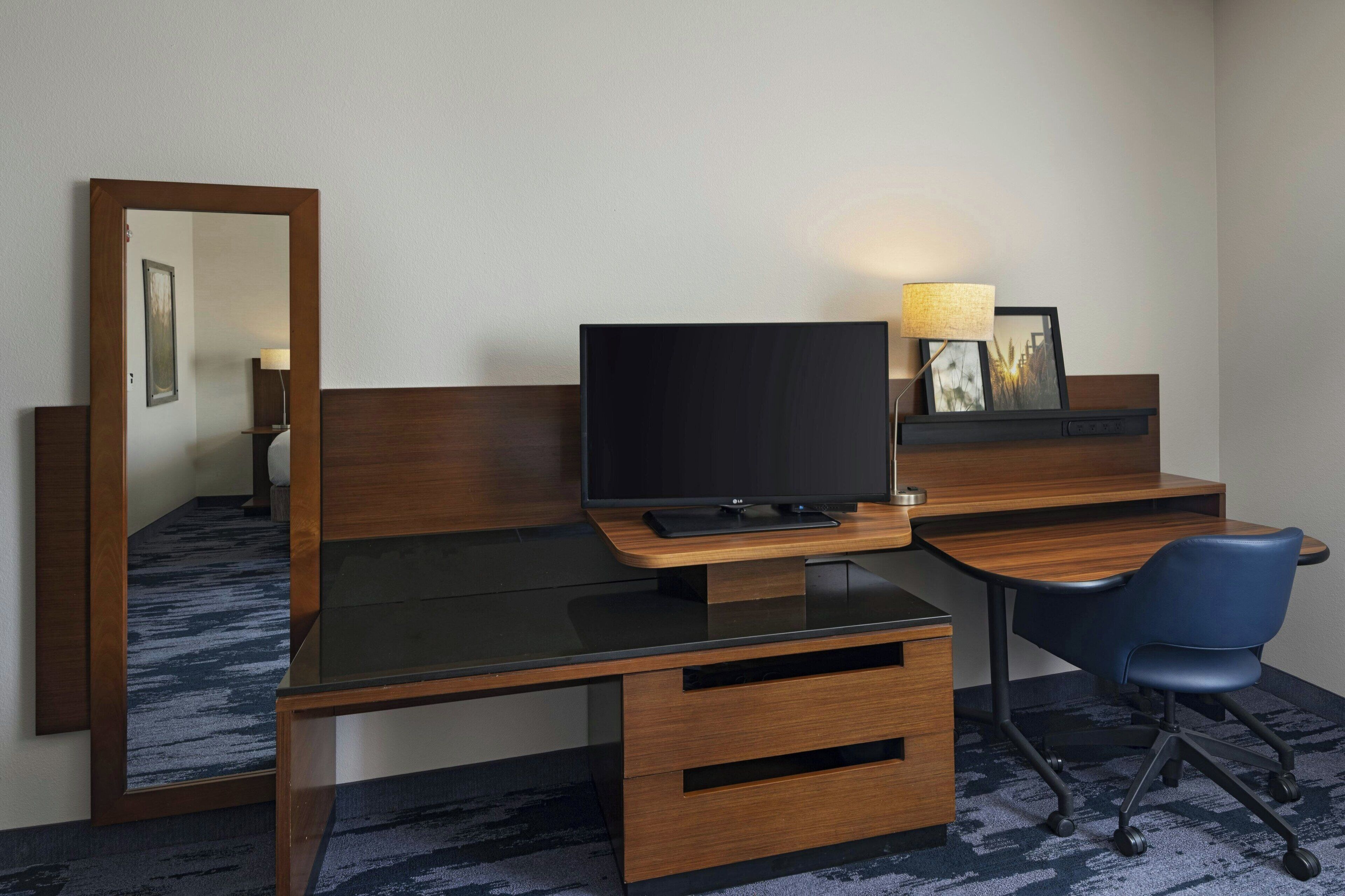 A hotel room with a wooden desk, a television, a chair, a mirror, and a lamp.
