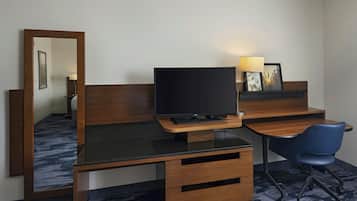 A hotel room with a wooden desk, a television, a chair, a mirror, and a lamp.