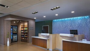 A hotel reception area with a self-service kiosk, a refrigerator stocked with snacks, and a sign indicating a corner market. - Fairfield Inn & Suites Des Moines Urbandale (Urbandale)