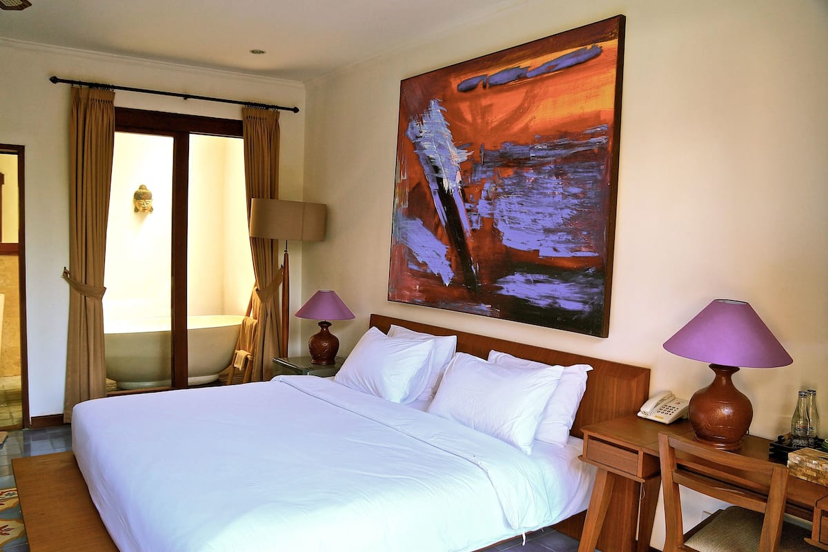 Habitación de hotel con una cama grande, dos lámparas de noche, un cuadro en la pared y un baño visible a través de una puerta de cristal.