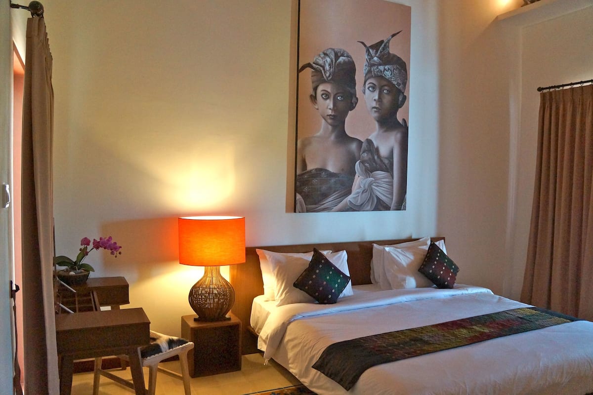 Una habitación de hotel con una cama, una mesita de noche con lámpara, una silla, una pequeña mesa y un cuadro colgado en la pared.
