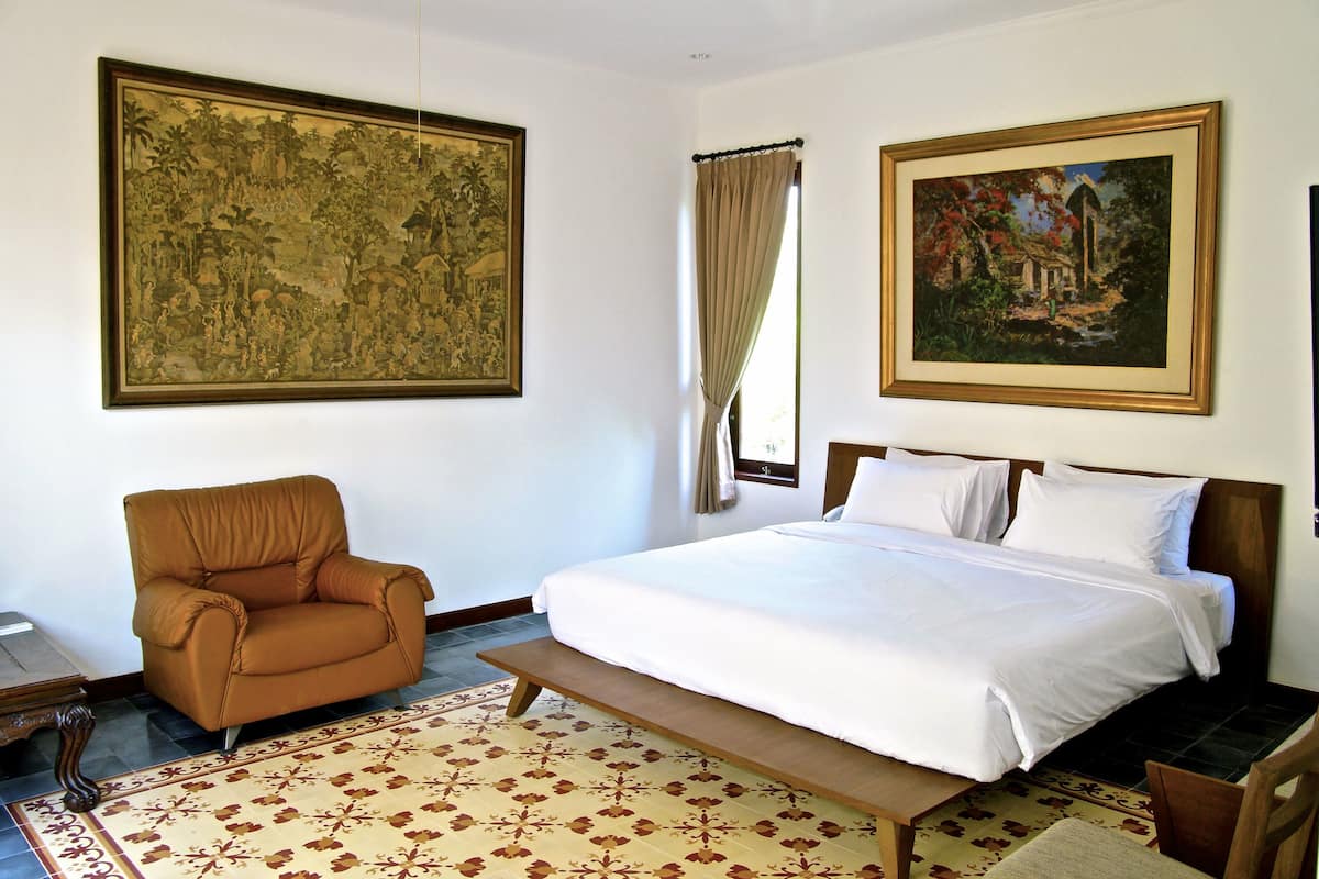 Habitación de hotel con una cama grande, un sillón marrón, un banco de madera, un tapete estampado y dos cuadros enmarcados en las paredes.
