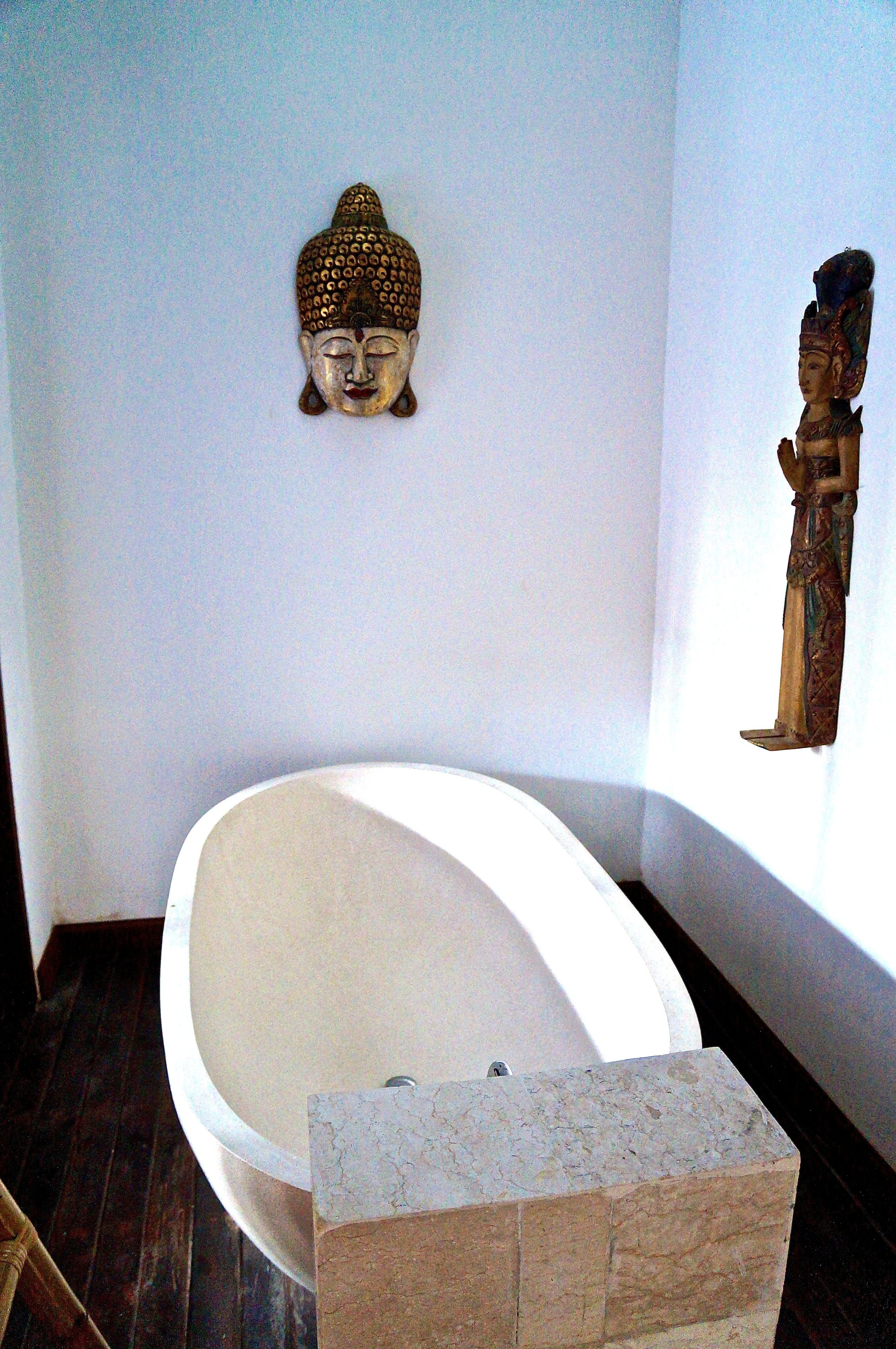 Un baño con una bañera independiente, una escultura de cabeza de Buda en la pared y una estatua a la derecha.