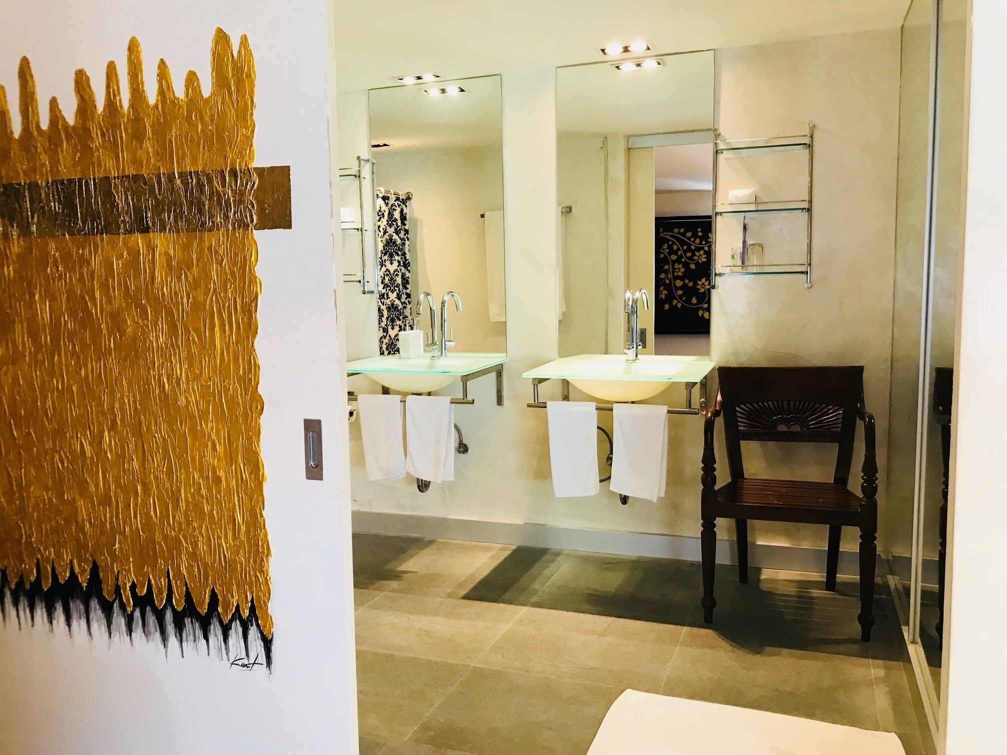 Un baño con un lavamanos de vidrio, dos espejos y una silla de madera.