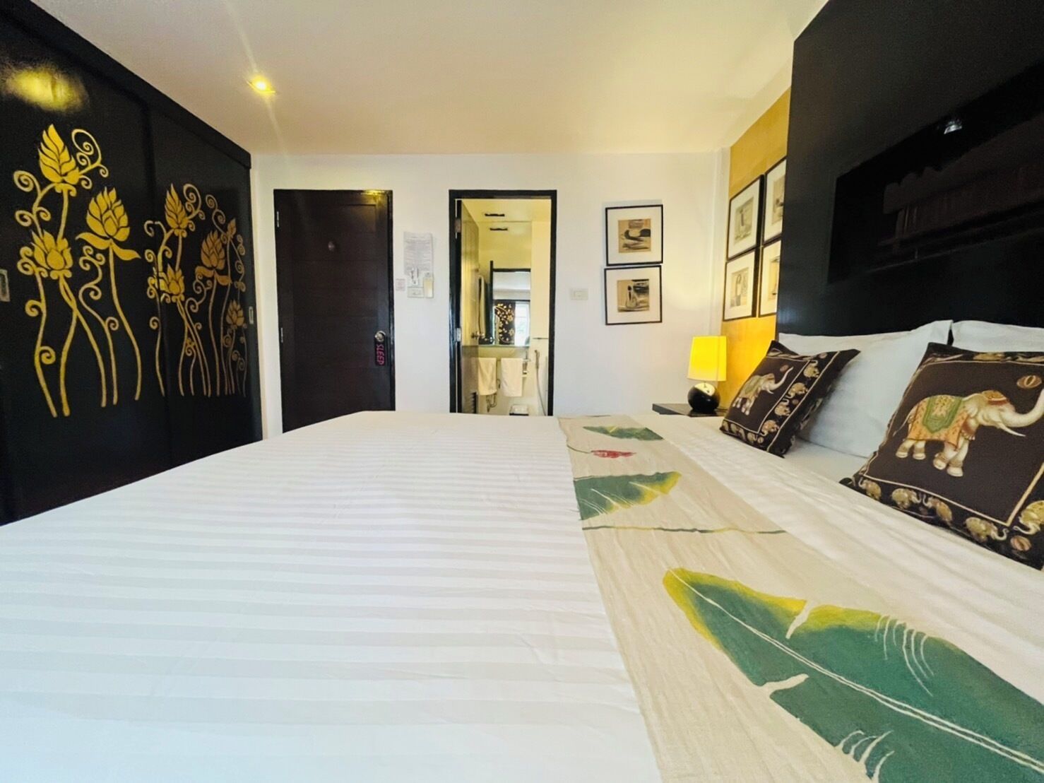A hotel room with a large bed, a black wall with yellow floral designs, a bedside table with a lamp, and a framed picture on the wall.