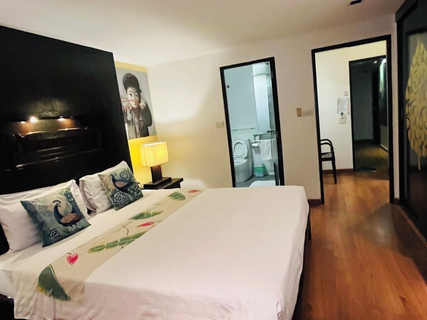 A hotel room with a large bed, a nightstand with a lamp, a painting on the wall, and a bathroom visible through an open door.