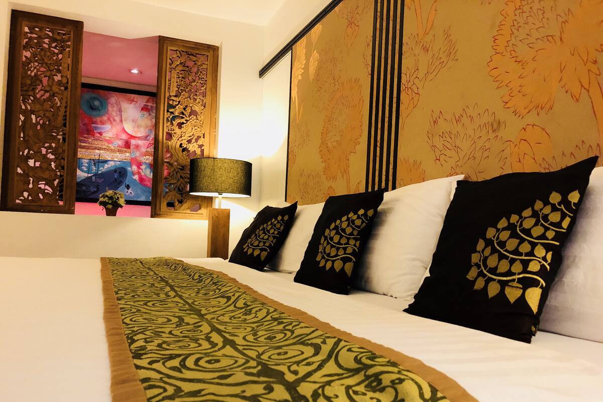 A hotel room with a large bed, patterned bedspread, decorative pillows, a nightstand with a lamp, and a wall with a floral tapestry.