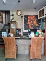 Un área de recepción con un mostrador, dos sillones de mimbre, un mural grande en la pared, una lámpara de techo y una escalera con una puerta de madera.