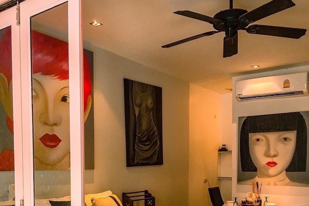 A bedroom with a bed, a desk, a chair, a ceiling fan, and artwork on the walls.