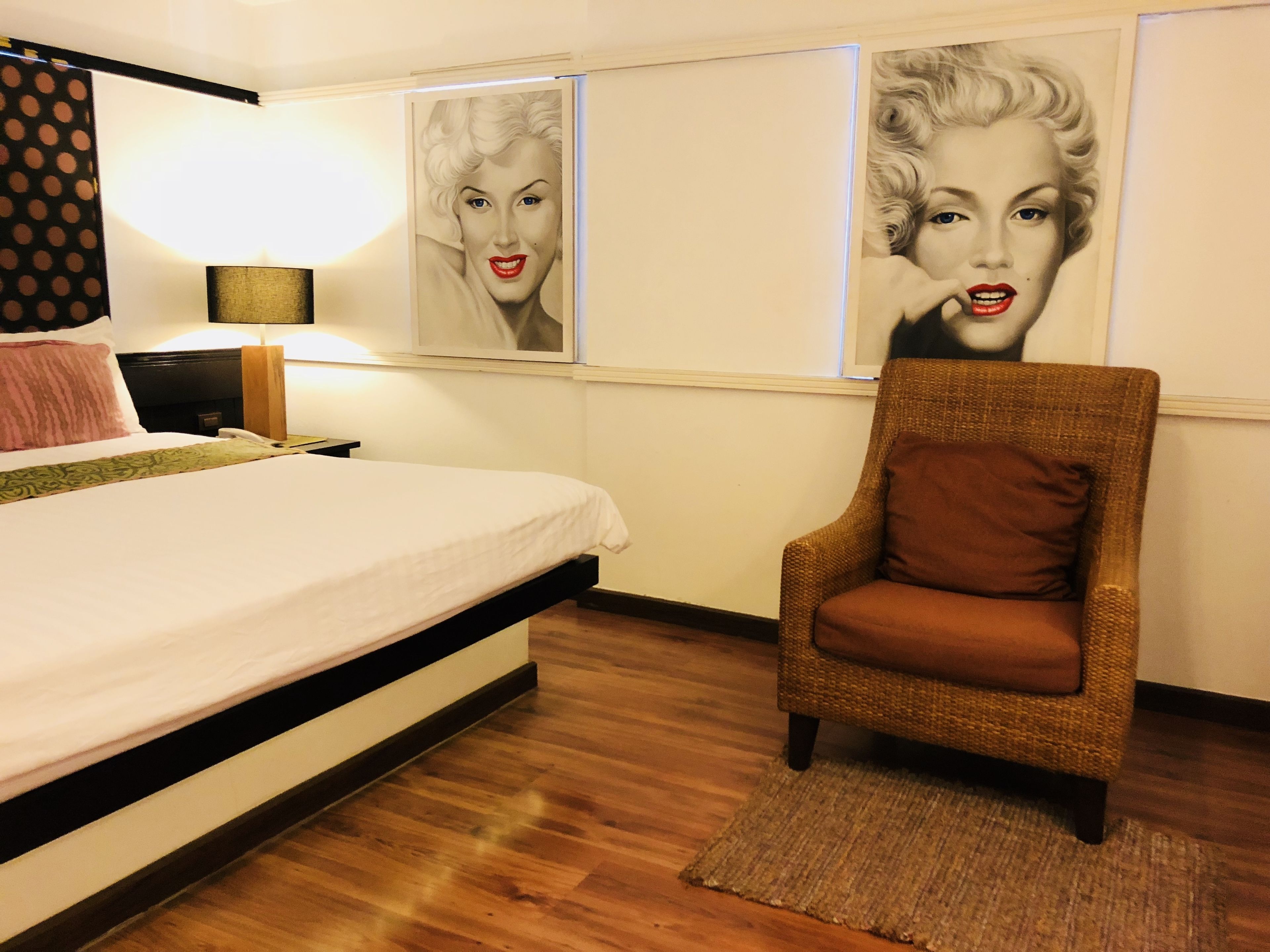 A hotel room with a bed, a chair, a lamp, and two posters on the wall.