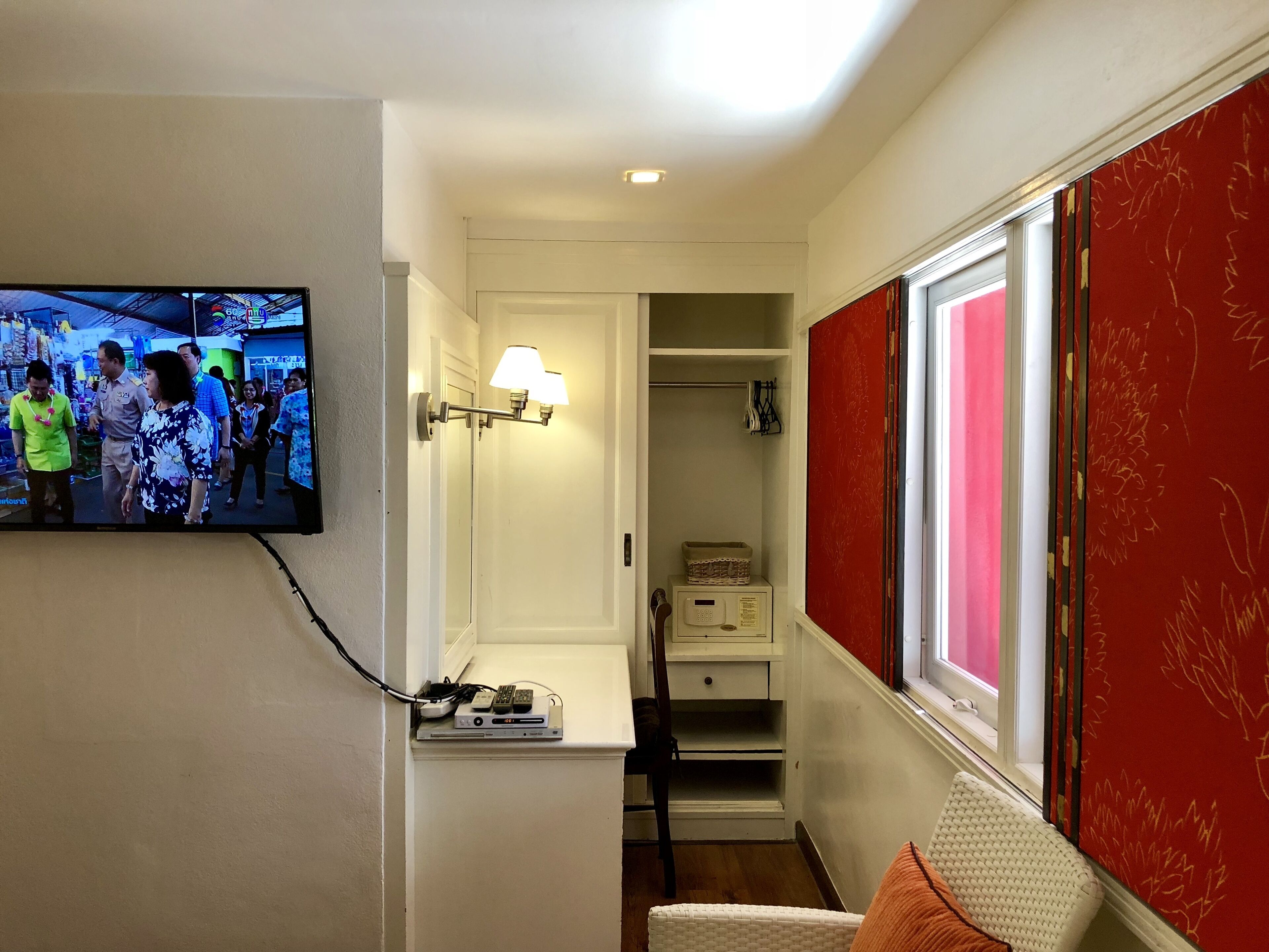 A hotel room with a television mounted on the wall, a bedside lamp, a small table with electronic devices, and a window with red curtains.