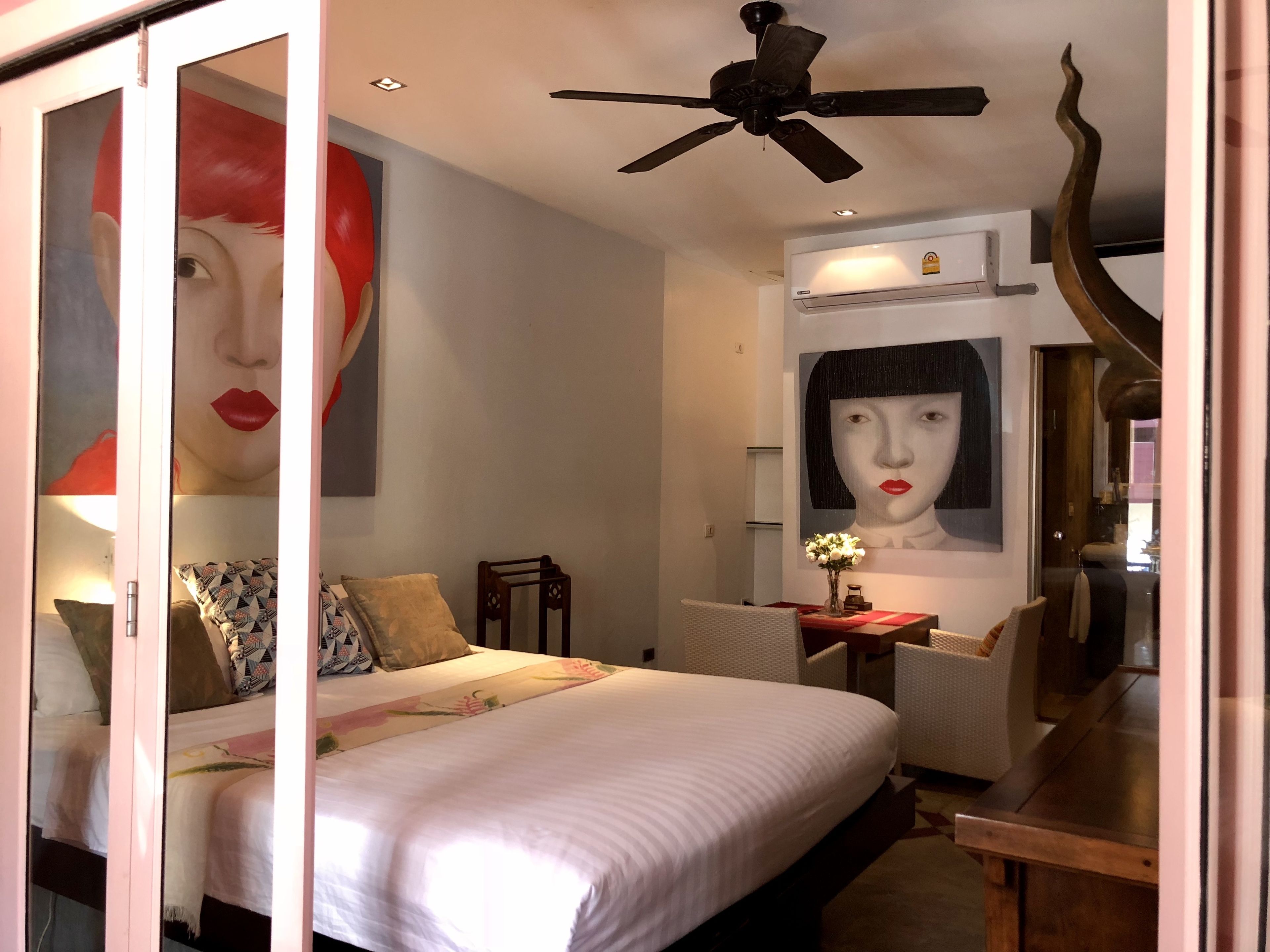 Une chambre à coucher comprenant un lit, un bureau, une chaise, un ventilateur de plafond et des œuvres d’art accrochées aux murs.