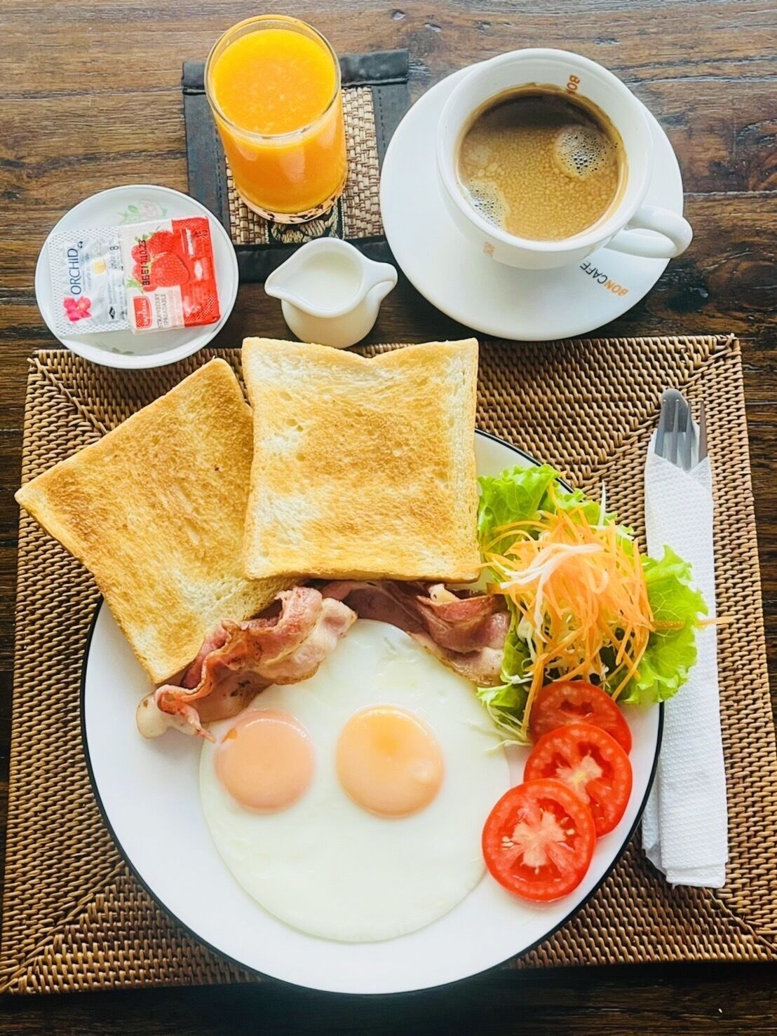 Un plato de desayuno con tostadas, huevos, tocino, lechuga y tomates, acompañado de un vaso de jugo de naranja y una taza de café.