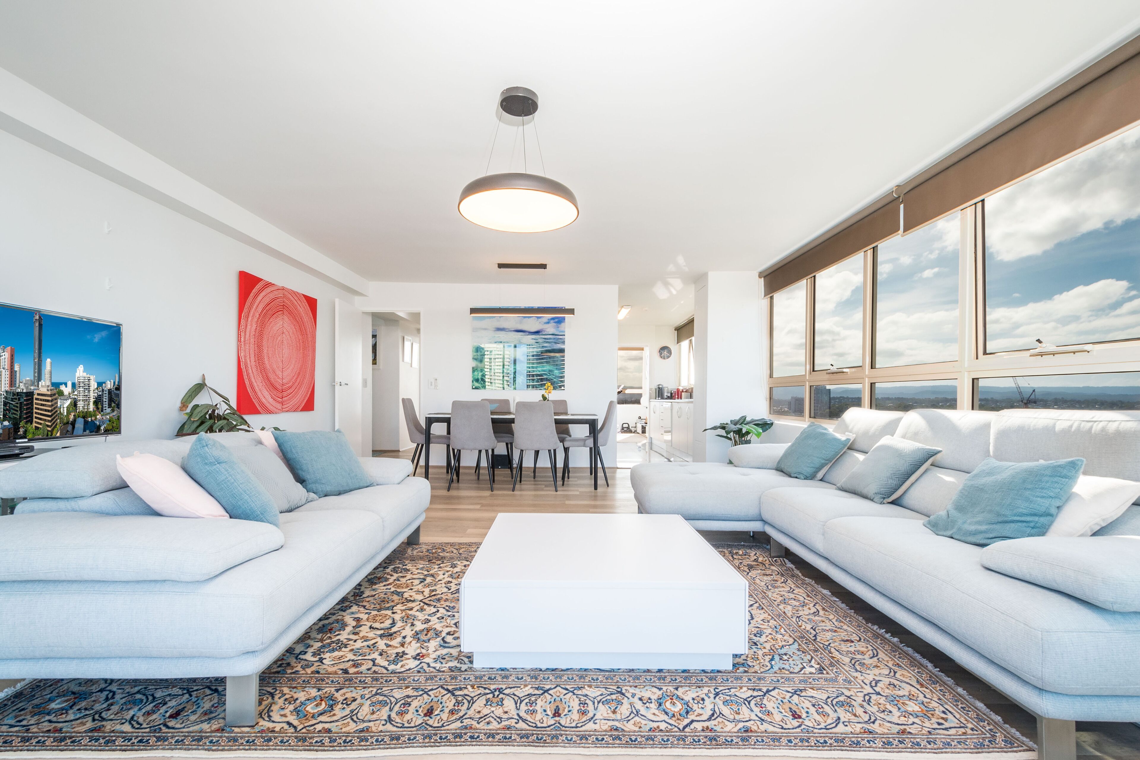 A modern living room with two white sofas, a coffee table, and a patterned rug. There is a dining area with a table and chairs, and a large window with a city view.