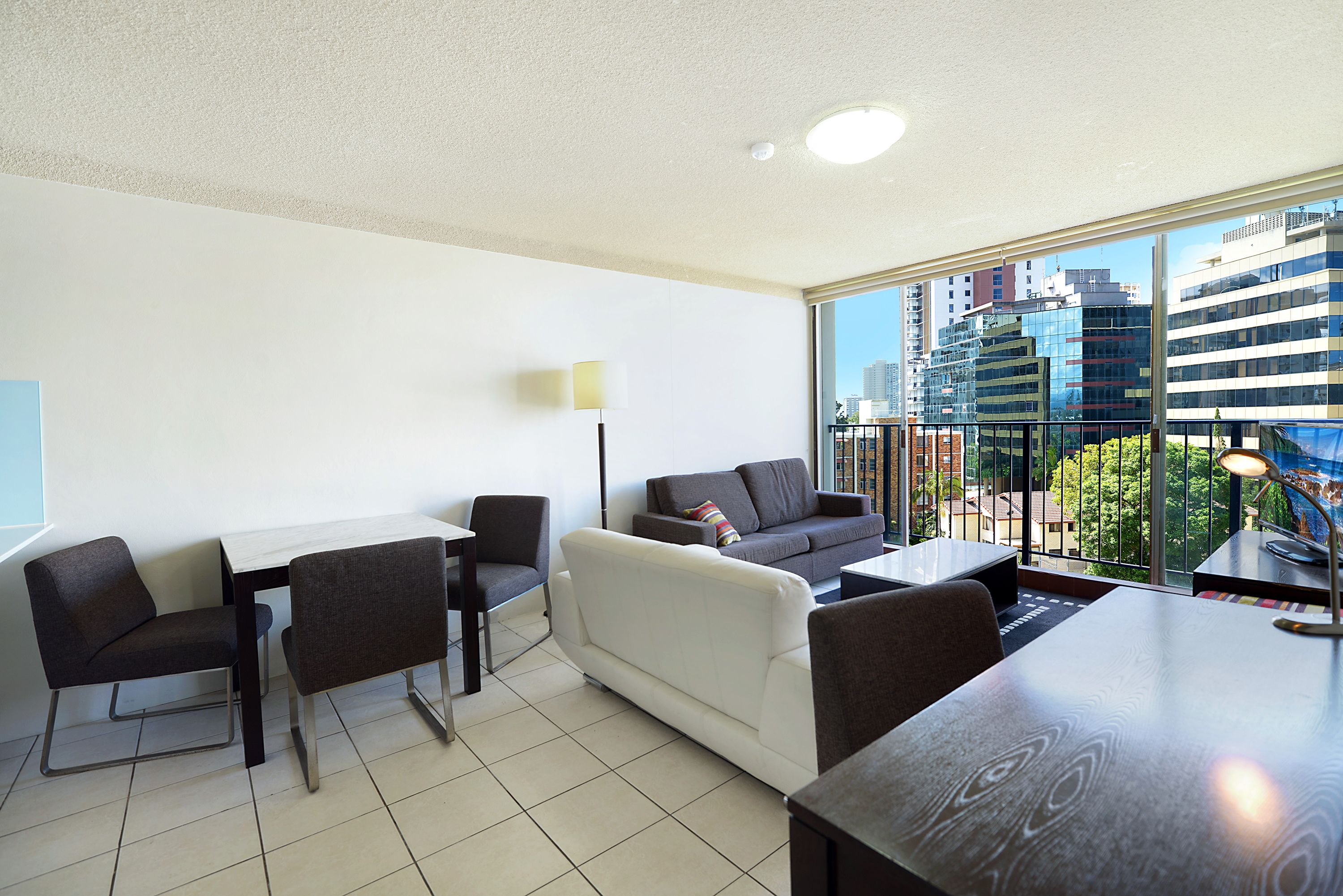 A modern living room with a white sofa, two armchairs, a dining table, and a large window offering a city view.