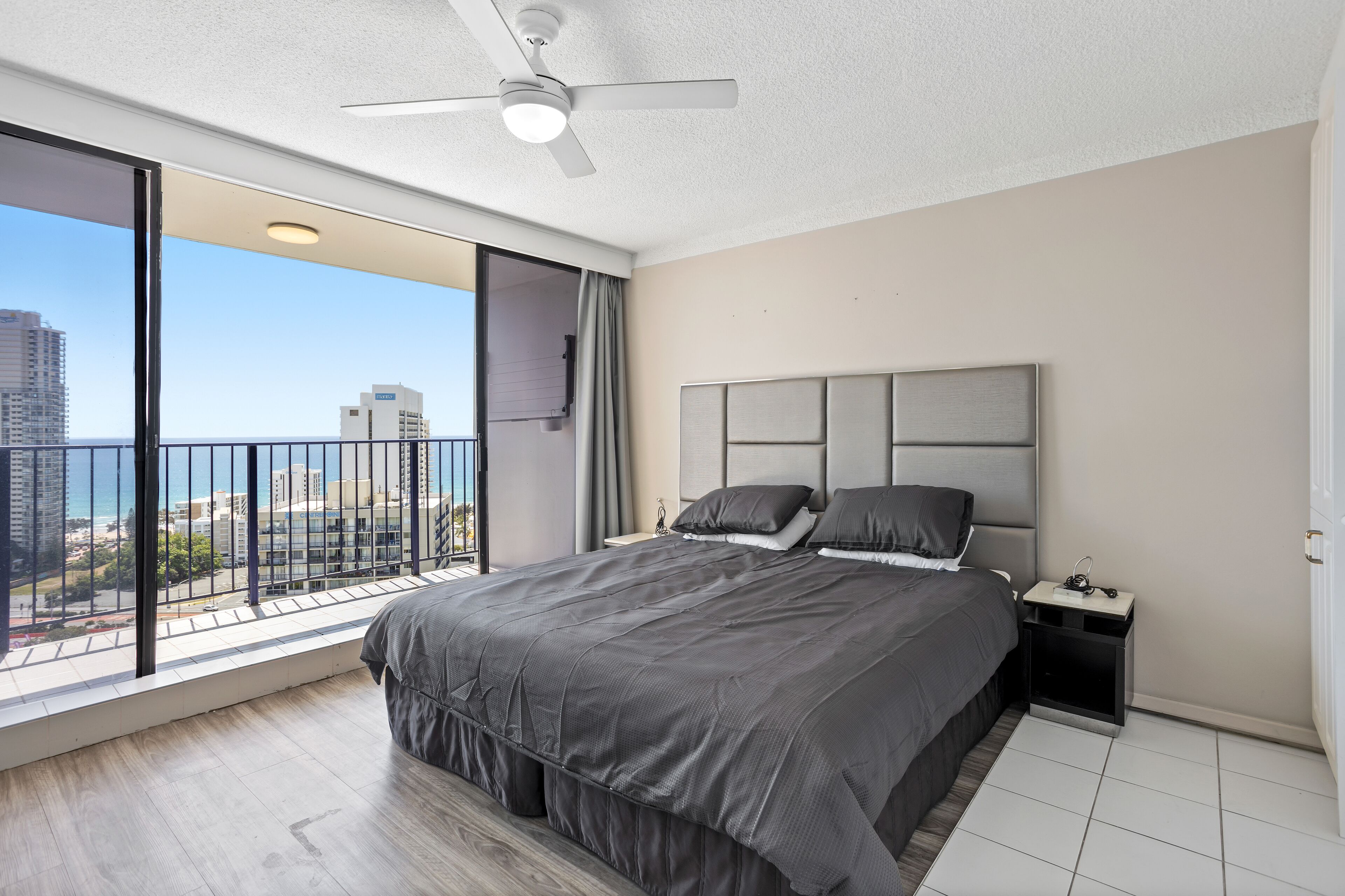 A modern bedroom with a large bed, a nightstand, a ceiling fan, and a balcony with a city view.