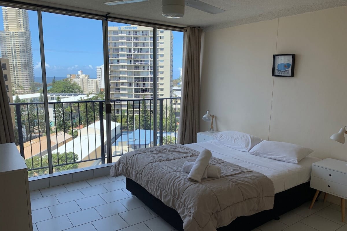 A hotel room with a large balcony, a bed with white linens, a bedside table with a lamp, and a framed picture on the wall.