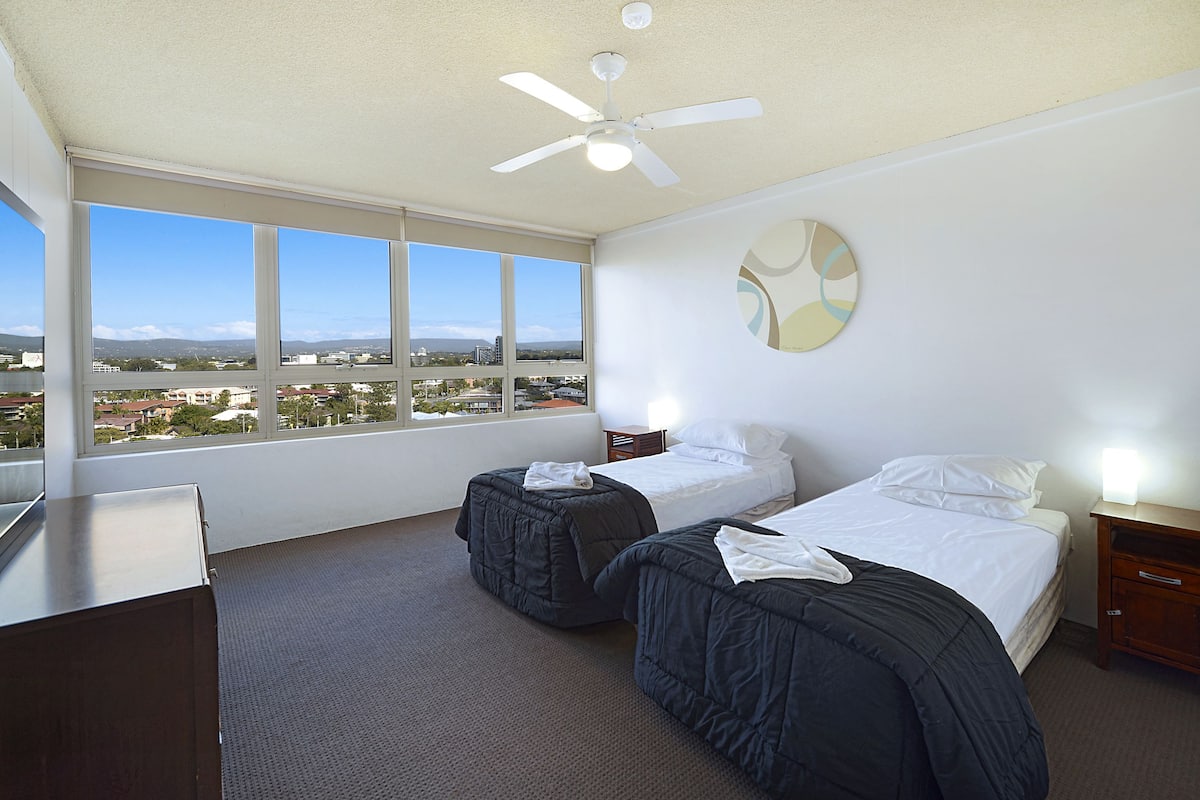 A hotel room with two beds, a ceiling fan, a large window with a city view, and a round wall decoration.